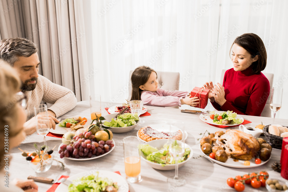 Warm-toned portrait of big happy family enjoying Christmas dinner together, focus on little girl giving present to mom