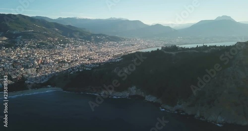 Ascending aerial drone footage of city, beach background pan right to reveal high key Alanya Castle cliff and Mediterranean Sea into the morning sun in Turkey. 4k at 23.97fps photo