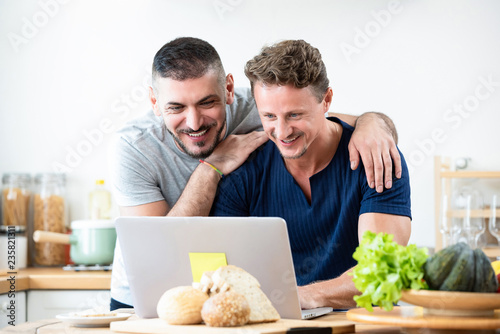 Happy gay male couple browsing internet together at home