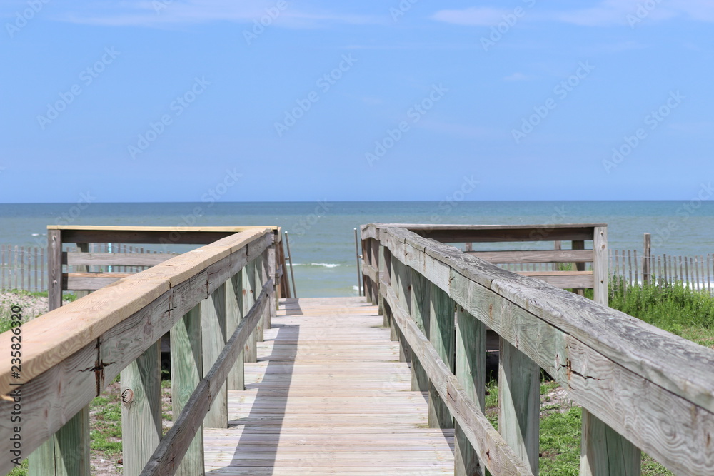 Perspective photograph of wood boardwalk railing beach access horizon blue sky, turquoise ocean and green grass.