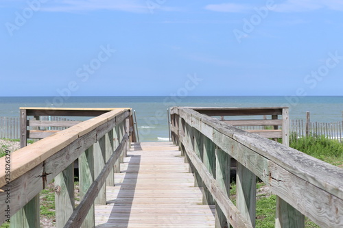 Perspective photograph of wood boardwalk railing beach access horizon blue sky  turquoise ocean and green grass.