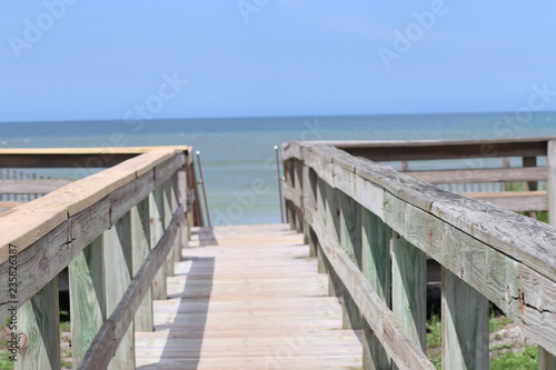 Perspective photograph of wood boardwalk railing beach access horizon blue sky  turquoise ocean and green grass.