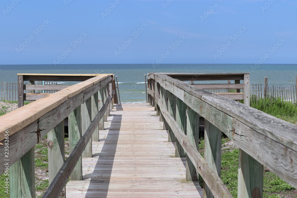Perspective photograph of wood boardwalk railing beach access horizon blue sky, turquoise ocean and green grass.