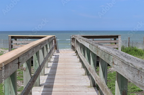 Perspective photograph of wood boardwalk railing beach access horizon blue sky, turquoise ocean and green grass. © Barb