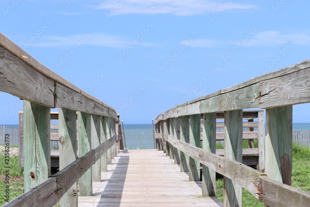 Perspective photograph of wood boardwalk railing beach access horizon blue sky, turquoise ocean and green grass. Perspective photograph of wood boardwalk railing beach access horizon blue sky, turquoise ocean and green grass.