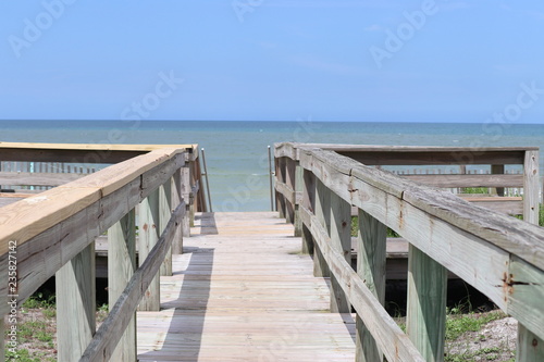Perspective photograph of wood boardwalk railing beach access horizon blue sky  turquoise ocean and green grass.