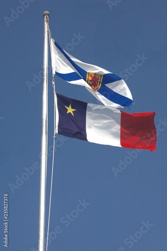 Chéticamp, Cape Breton, Nova Scotia, Canada: Flags of Nova Scotia (top) and Acadia (bottom), waving in the wind on flagpoles against a deep blue cloudless sky.