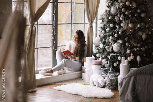 Nice dark-haired girl dressed in pants, sweater and warm slippers reads a book sitting on the windowsill of a panoramic window in the room next to the New Year tree, gifts and candles