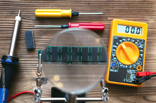 Retro memory module chip through magnifying glass mounted on the soldering stand and multimeter, screwdriver and soldering iron on technician engineer table background.
