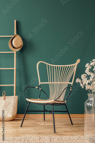Flowers next to rattan armchair in green living room interior with hat on ladder above bag. Real photo