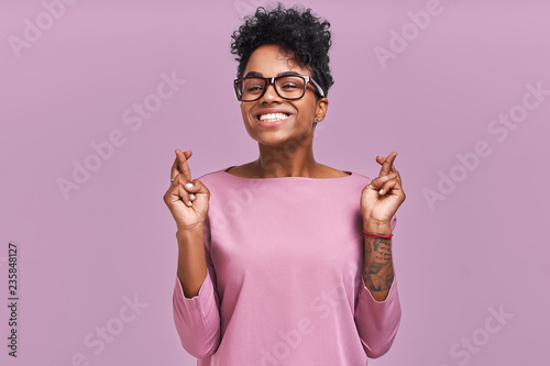 I have to win. Portrait of young female has dark skin, clenches teeth, raises fingers crossed, makes desirable wish, wears round spectacles and casual clothes, waits for good news