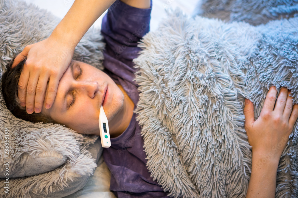 A young man lies under a blanket and clings to his forehead. Check the temperature of the thermometer. to close. A young man lies under a blanket and clings to his forehead. Check the temperature of the thermometer. to close.