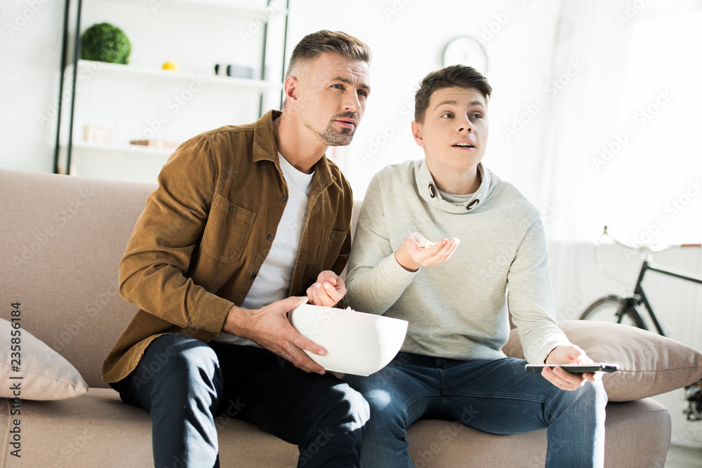 serious father and teen son watching tv and holding bowl of popcorn at home