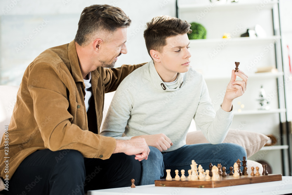 father hugging teen son while he looking at chess figure at home father hugging teen son while he looking at chess figure at home