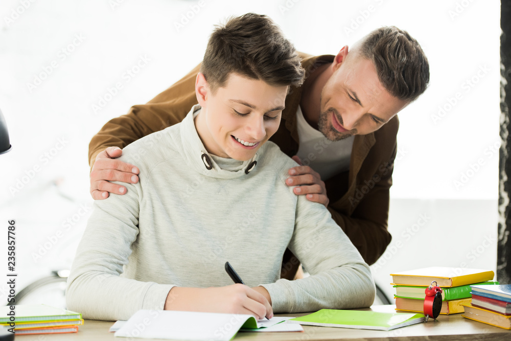 smiling father hugging teen son while he doing homework and writing something smiling father hugging teen son while he doing homework and writing something