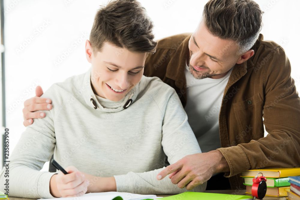 smiling father hugging cheerful teen son while he doing homework smiling father hugging cheerful teen son while he doing homework
