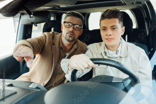 handsome father pointing on something while teaching teen son driving car