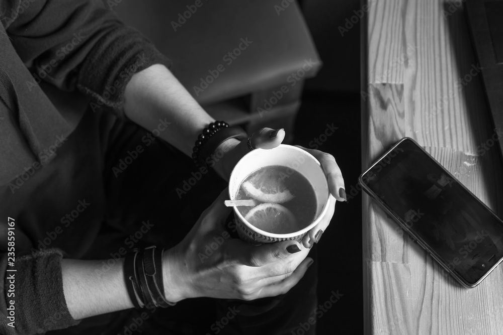 Top view. Student woman holds with two hands a hot cup of tea with sea buckthorn Hippophae and a slice of orange. Workplace, notebook with pen and laptop, mobile phone, smartphone, black and white
