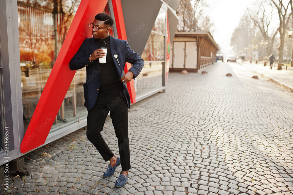Amazingly looking african american man wear at blue blazer with brooch, black turtleneck and glasses posed at street. Fashionable black guy with cup of coffee.