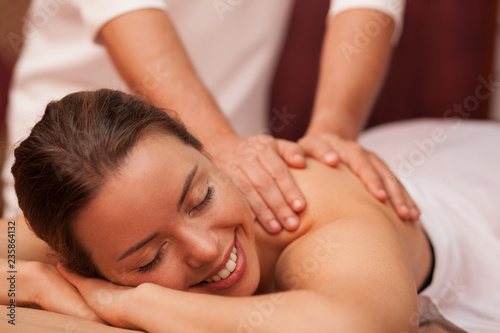 Attractive joyful young woman smiling cheerfully why professional masseur massaging her shoulder. Gorgeous woman relaxing at day spa. Therapist giving soothing massage to female client. Beauty concept