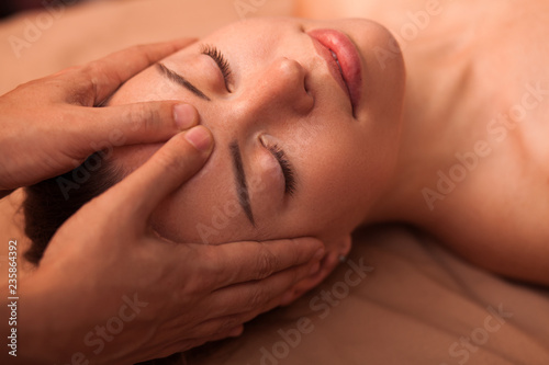 Close up of a happy beautiful woman smiling with her eyes closed, receiving soothing head massage. Young attractive woman enjoying face massage at spa center. Skincare, health, pampering concept