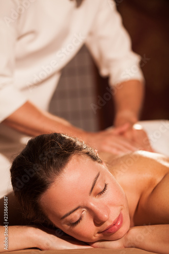 Vertical shot of a beautiful woman relaxing at spa center, receiving back massage. Attractive female enjoying soothing massage therapy. Professional masseur working. Resort, hotel, recreation concept.