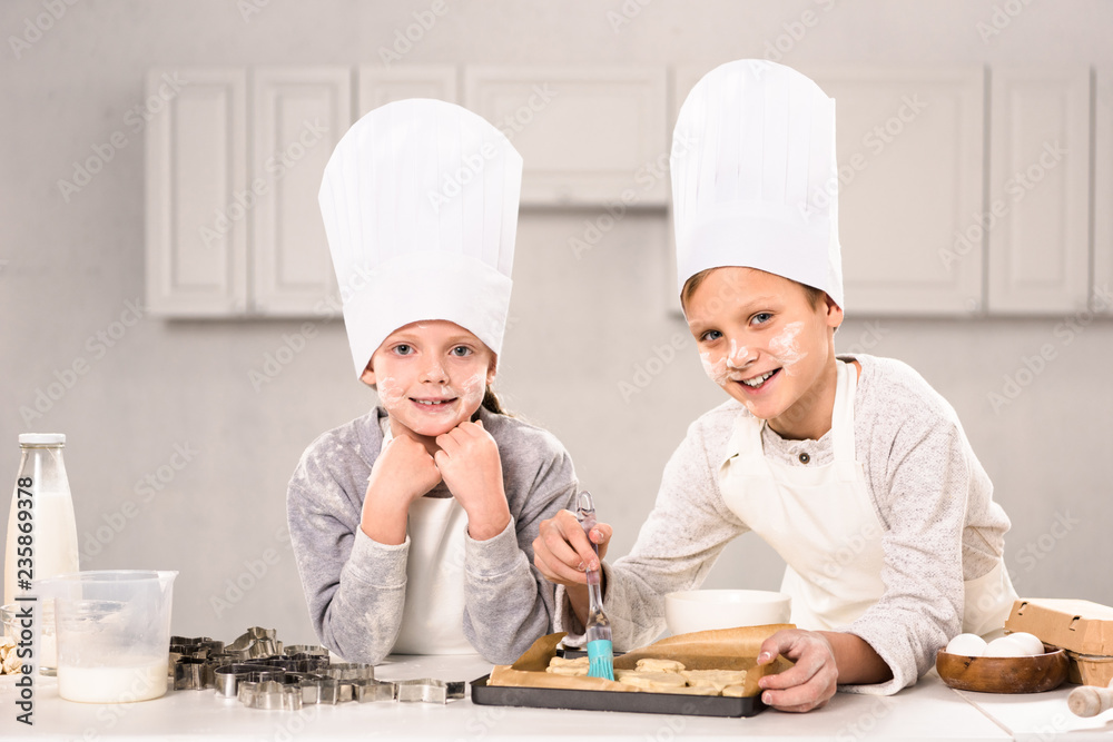 joyful children in aprons brushing cookies on baking tray in kitchen