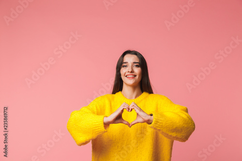 Beautiful young emotional woman posing isolated over pink background showing heart gesture.