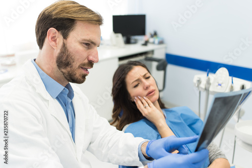 Woman holding her hurtful tooth having painful face expression sitting at the dentist office while beautiful young successful dentist is holding and looking at the teeth x-ray.