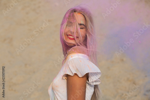 Closeup shot of emotional blonde girl with red lipstick posing in a cloud of pink dry paint Holi at the desert