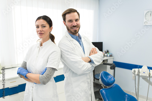 Young beautiful successful team of two smiling dentists working in modern dental clinic. Dentist and his assistant crossed hands looking at camera and smiling. Dentistry and doctors concept.