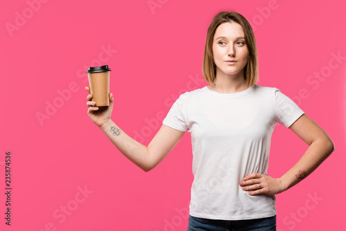 pretty tattooed girl showing disposable cup of coffee isolated on pink