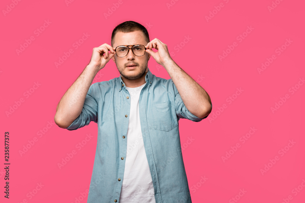 serious young man adjusting eyeglasses and looking at camera isolated on pink serious young man adjusting eyeglasses and looking at camera isolated on pink