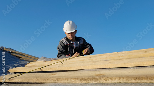 Adult professional builder in hardhat measuring length of wood lumber on site under blue sky