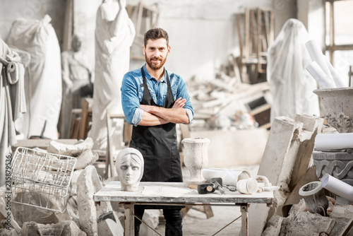 Portrait of a handsome sculptor in blue t-shirt and apron working with stone sculptures on the table at the old atmospheric studio