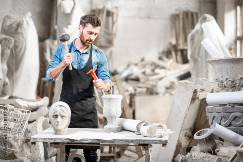 Fototapeta Portrait of a handsome sculptor in blue t-shirt and apron working with stone scu