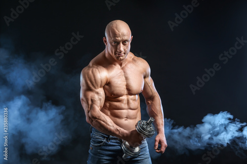 Muscular bodybuilder with dumbbell in smoke on dark background