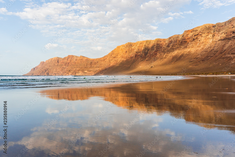 Mirror landscape with volcanic mountains and blue cloudy sky reflected in the wet sand. Famara beach at sunset, Lanzarote, Canary Islands, Spain.