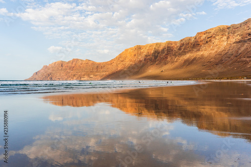 Mirror landscape with volcanic mountains and blue cloudy sky reflected in the wet sand. Famara beach at sunset, Lanzarote, Canary Islands, Spain.
