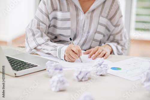 Faceless shot of woman working in office and writing document at table with crumpled papers around