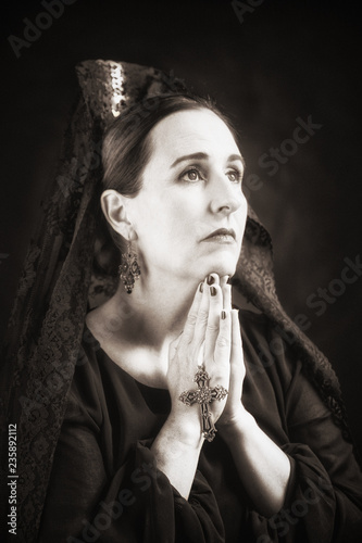 Spanish lady with devoted look, interlocking hands with cross praying, looking up to the light wearing traditional clothing: black veil / mantilla and a high comb / peineta. photo
