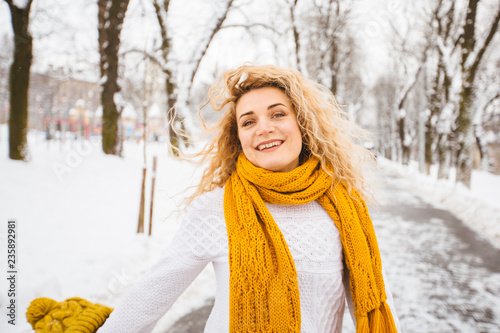 Blond curly playful hipster woman on street, ljumping, enjoying winter smiling at snowy park. Model wearing white sweater, yelow knitted hat, sarf, gloves. City lifestyle concept. photo