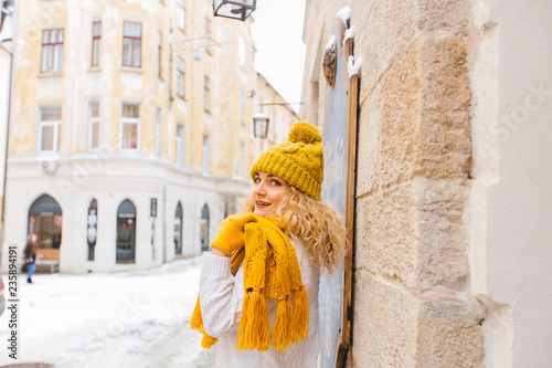 Blond curly romantic hipster woman walking on the city street, enjoying winter smiling at snowy day. Female wearing white sweater, yelow knitted hat, sarf, gloves. City lifestyle concept.