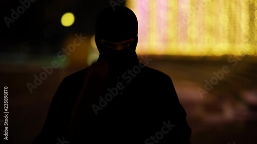 A man dressed in dark clothes and a balaclava goes with a bat in an abandoned park. There is an early winter outside and garlands hang around. His face is not visible in backlight. He is set up to fig