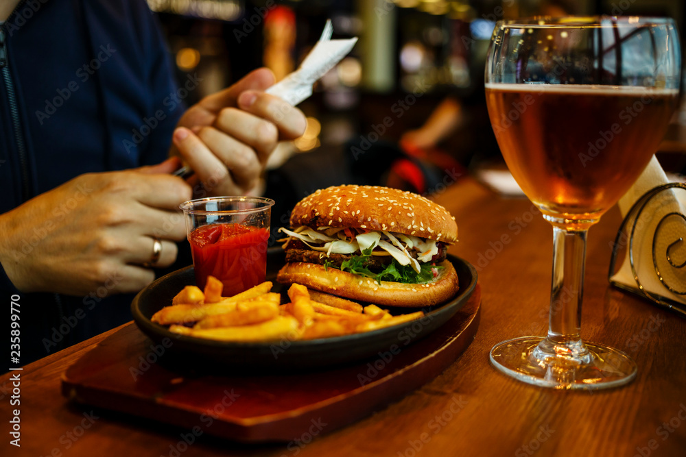 Burger with French fries and Beer, fork and knife in male hands on wooden table background Burger with French fries and Beer, fork and knife in male hands on wooden table background