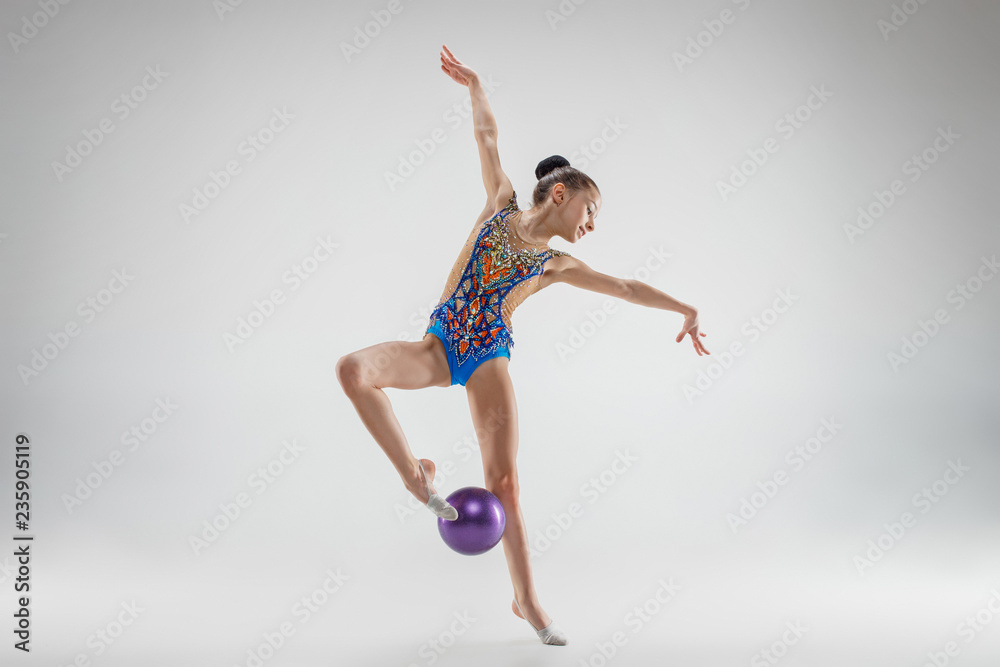 Fototapeta premium The teen female little girl doing gymnastics exercises with ball on a gray studio background. The gymnastic, stretch, fitness, lifestyle, training, sport concept