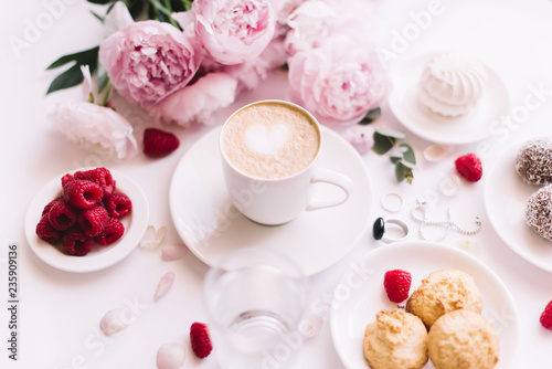 Fototapeta Naklejka Na Ścianę i Meble -  Delicious morning breakfast table essentials: fresh cappuccino coffee, marshmallow, cookies, chocolate, red raspberries, glass of water and tender pink blossoming peony bouquet on the white background