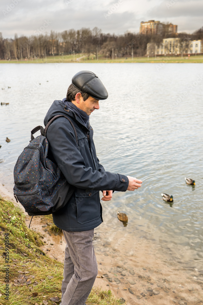 Handsome dark-haired middle-aged man in jacket, cap and backpack feeds ducks on pond in city park in late autumn. Authentic lifestyle moments Handsome dark-haired middle-aged man in jacket, cap and backpack feeds ducks on pond in city park in late autumn. Authentic lifestyle moments