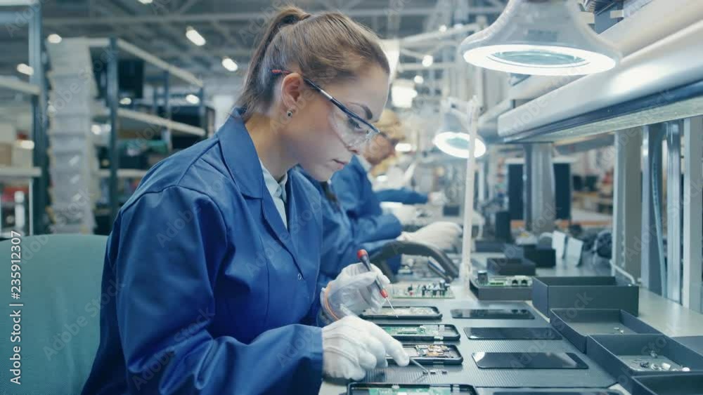 Woman Electronics Factory Worker in Blue Work Coat and Protective Glasses is Assembling Smartphones with Tweezers and Screwdriver. High Tech Factory Facility with more Employees in the Background. 