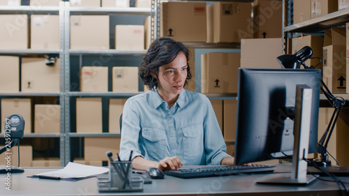 Fotografía Female Inventory Manager Works on a Computer while Sitting at Her Desk, Marking Orders in Clipboard Checklist